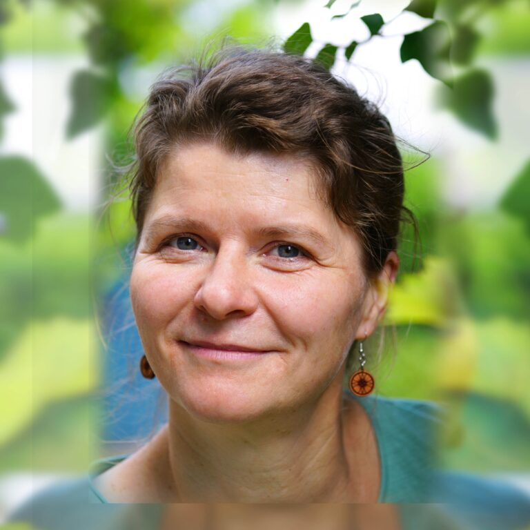 Eine Frau mittleren Alters mit hellbraunen Haaren und blauen Augen lächelt im Freien; sie trägt Ohrringe aus Holz und ein blaugrünes Oberteil und ist von grünem Laub umgeben.