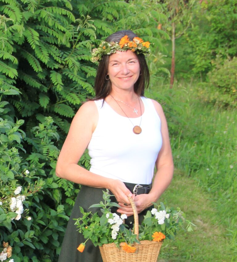 Eine Frau in einem weißen Oberteil und mit einem Blumenkranz im Haar steht im Freien, lächelt und hält einen Weidenkorb voller Blumen und Grünpflanzen in der Hand. Im Hintergrund sind üppig grüne Pflanzen zu sehen.