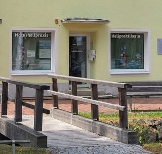 Ein kleines gelbes Gebäude mit zwei Schildern in deutscher Sprache an den Fenstern – „Naturheilpraxis“ und „Heilpraktikerin“ – sowie einer Holzrampe, die zur Eingangstür führt.