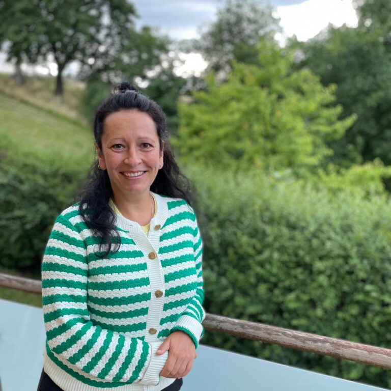 Eine Frau mit langen dunklen Haaren, die einen grün-weiß gestreiften Pullover trägt, steht auf einem Balkon vor dem Hintergrund üppiger Vegetation und Bäume und lächelt in die Kamera.