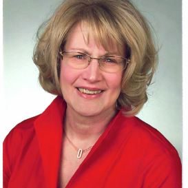Lächelnde Frau mit kurzen blonden Haaren, die eine Brille und eine leuchtend rote Bluse trägt und vor einem schlichten hellgrauen Hintergrund posiert.